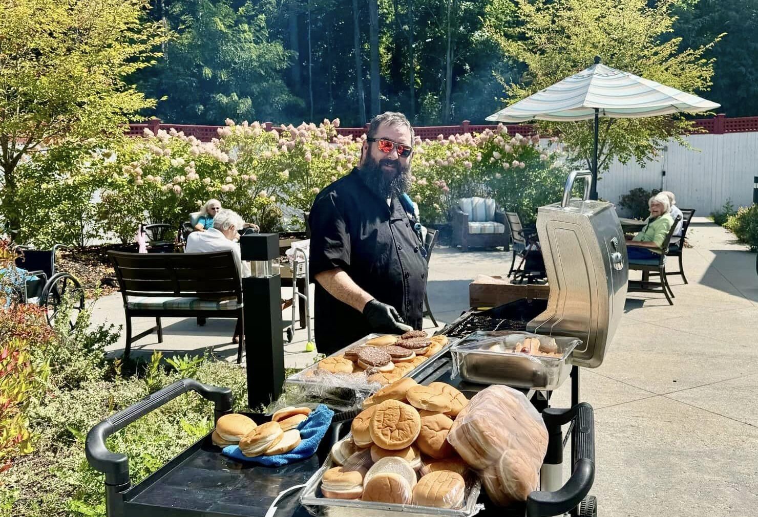A bearded man smiles while cooking burgers on a grill as several older people sit on patio furniture in the background.