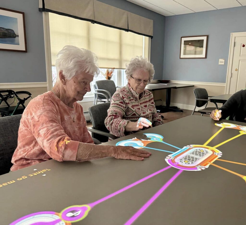 Two older women sit at a Tovertafel table and play an interactive light-projected game.