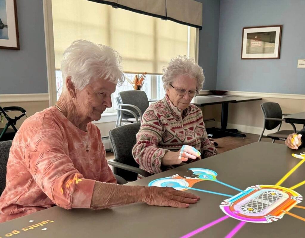 Two older women sit at a Tovertafel table and play an interactive light-projected game.